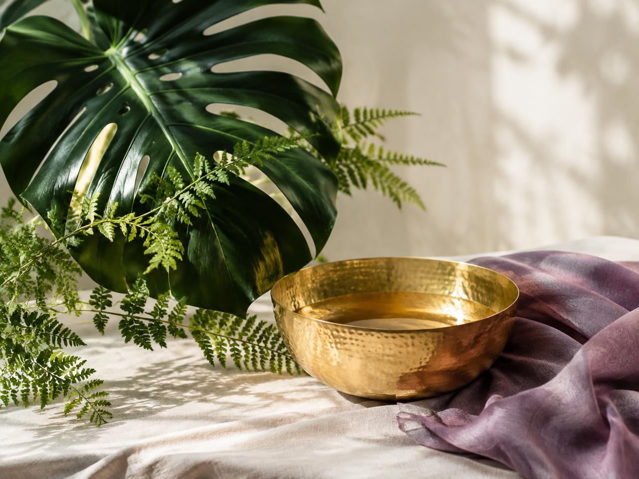 A gold bowl of warm oil beside fern and monstera leaves and amethyst silk